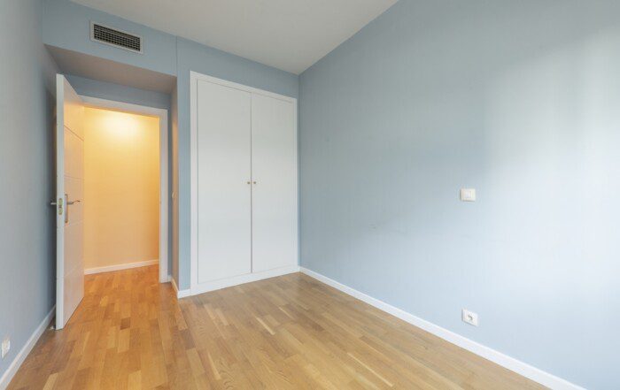 Bedroom of an empty house with laminated oak flooring with a white built-in wardrobe, ducted air conditioning and white carpentry.