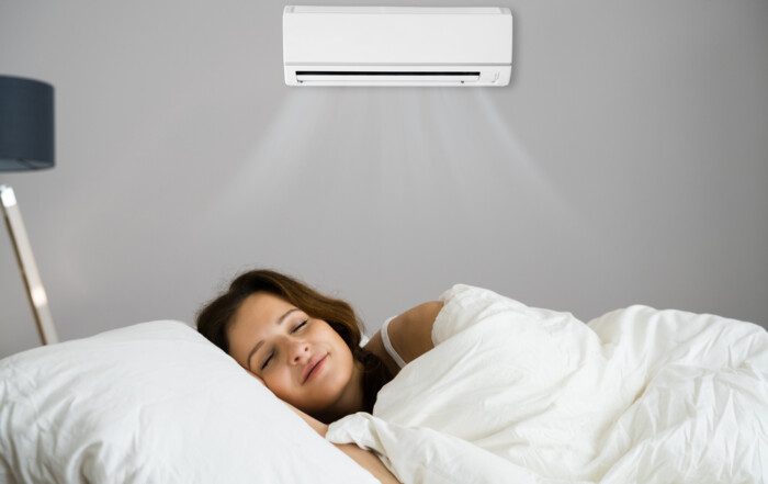 A woman asleep in bed, wrapped in a white blanket, with an air conditioner providing a cool and comfortable environment.